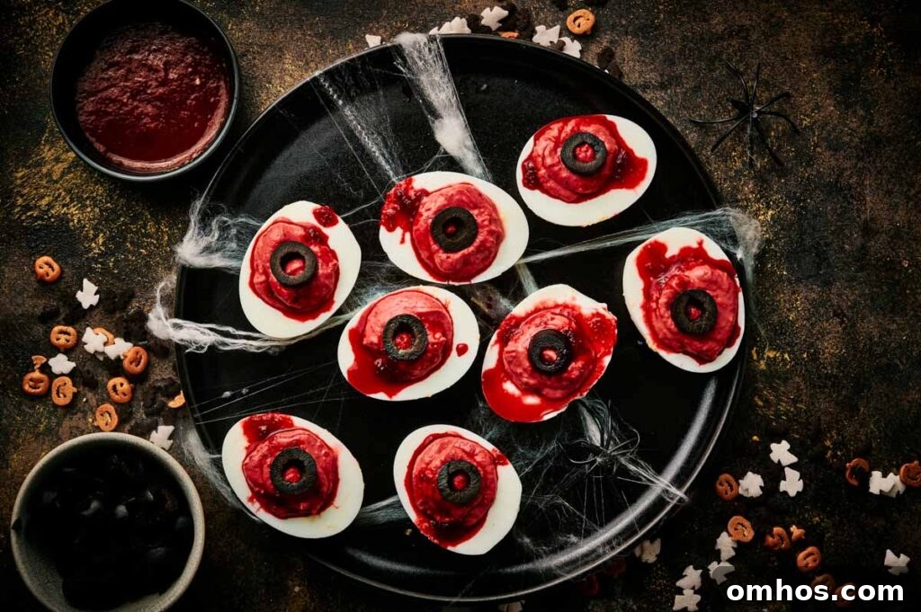 A black tray holds eight halloween deviled eggs decorated to look like bloody eyeballs, surrounded by fake spider webs and Halloween-themed confetti.