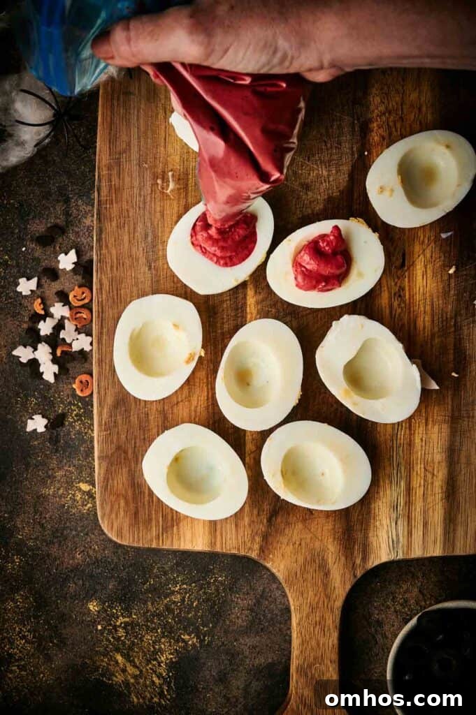 A hand expertly fills hollowed hard-boiled egg halves with a red filling using a piping bag on a wooden board, crafting spooky Halloween deviled eggs. Eerie decorations are scattered nearby, perfecting the festive atmosphere.