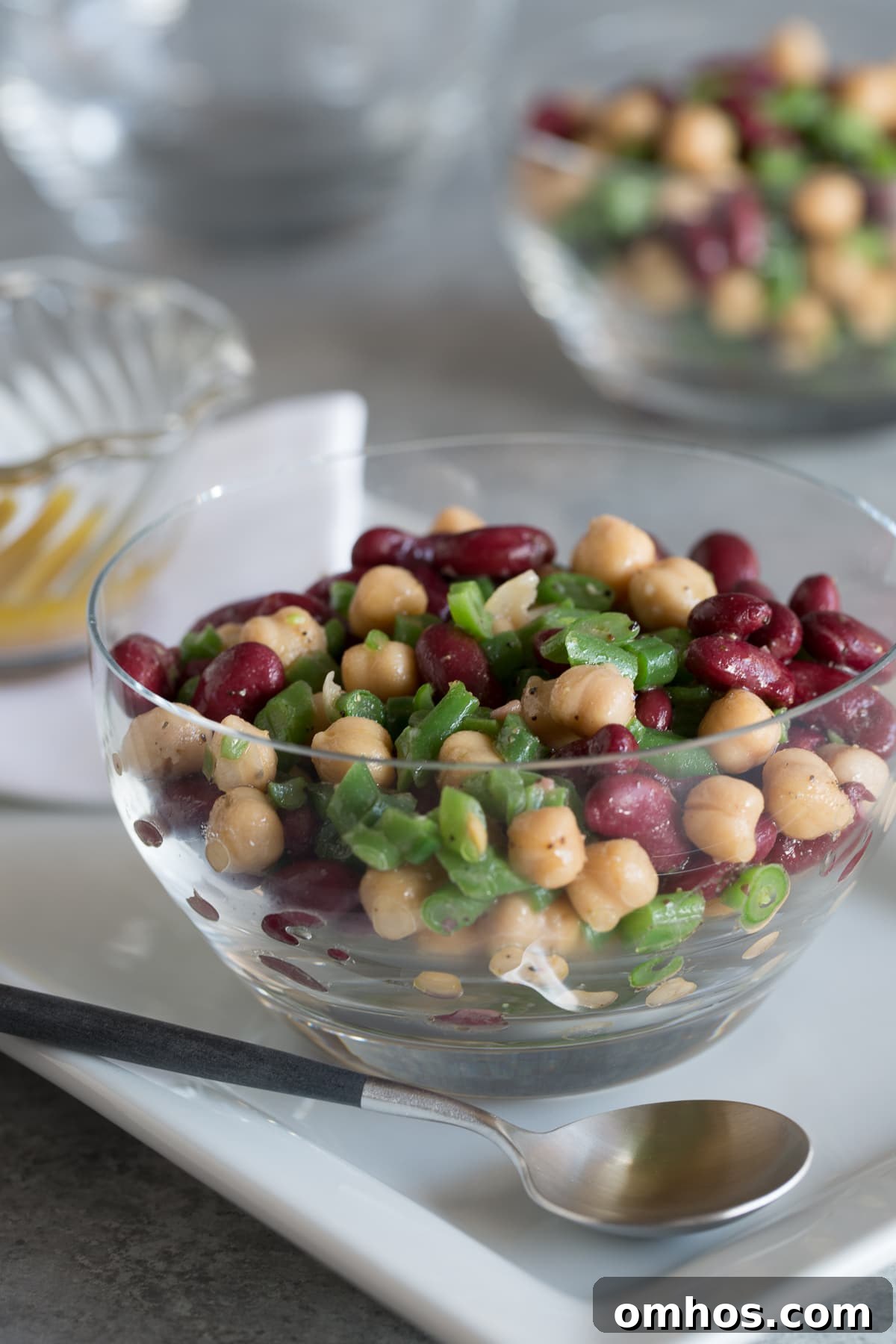 Colorful 3-bean salad in a glass bowl with a silver spoon and vinaigrette.