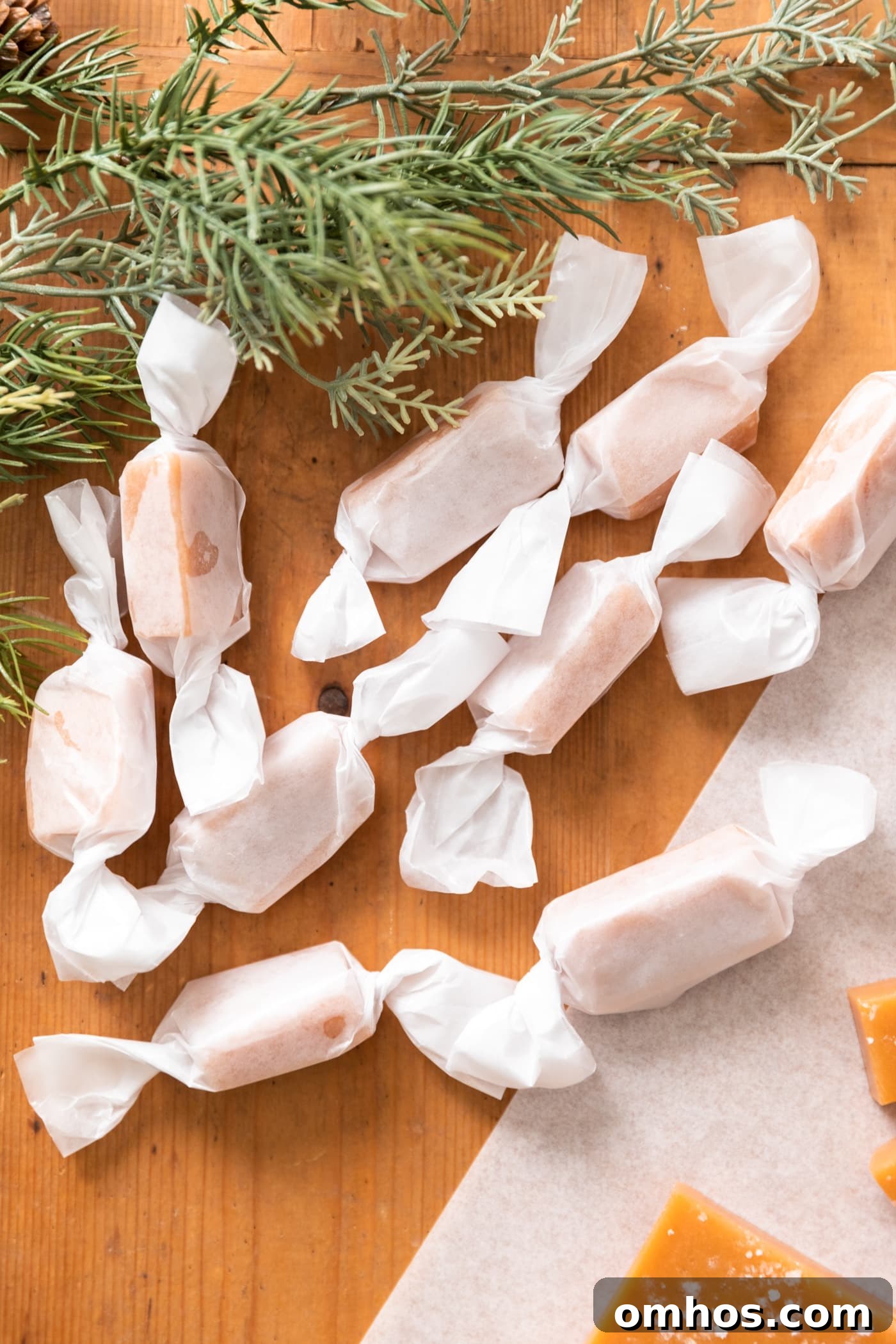 Wax wrapped pieces of caramel sitting on wood surface with greenery in background and unwrapped caramels beside
