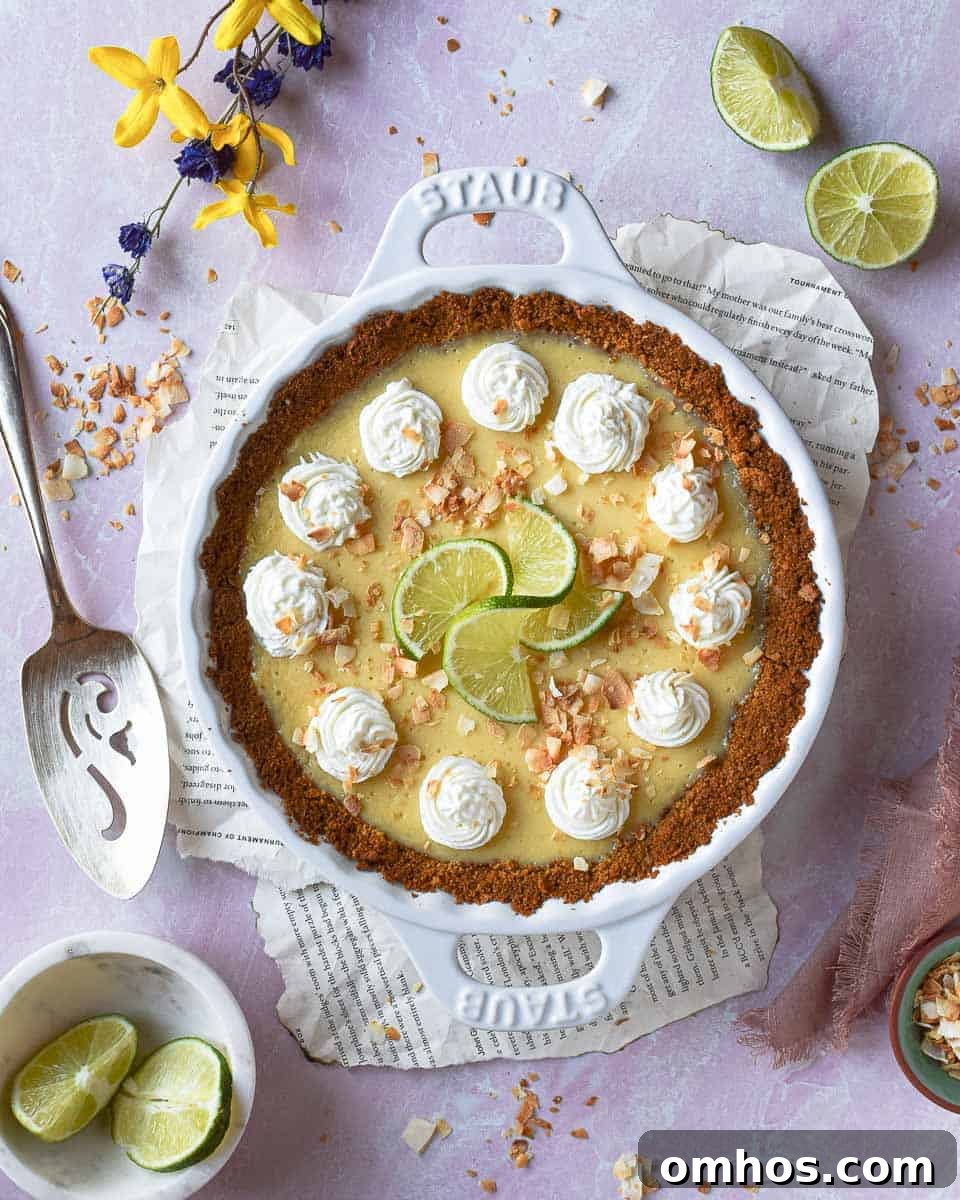 Overhead shot of coconut key lime pie in white pie dish placed on a pink background. Scattered limes in the background.