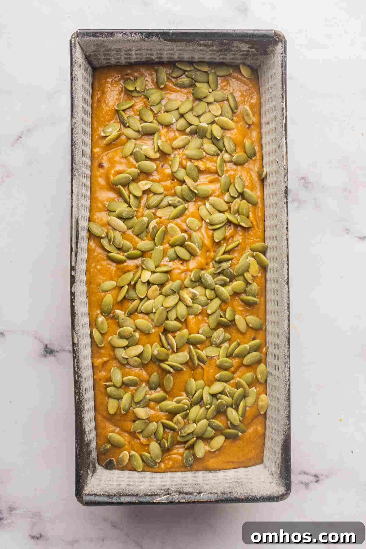 Starbucks-Style Pumpkin Bread Recipe 9 A loaf pan with batter topped with raw pumpkin seeds, ready to be baked.