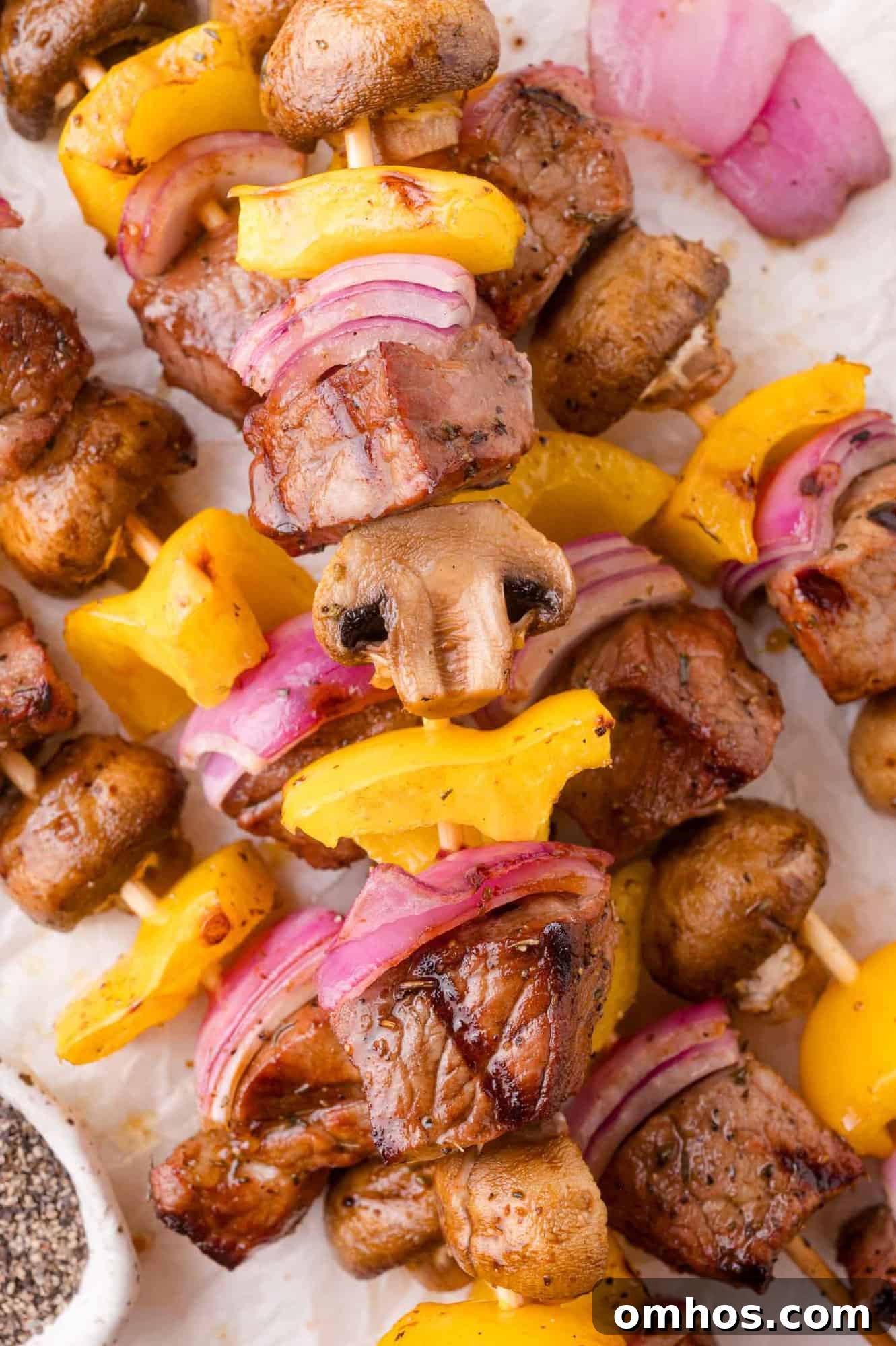 Grilled skewers with chunks of steak, yellow bell peppers, red onions, and mushrooms arranged on a white surface.