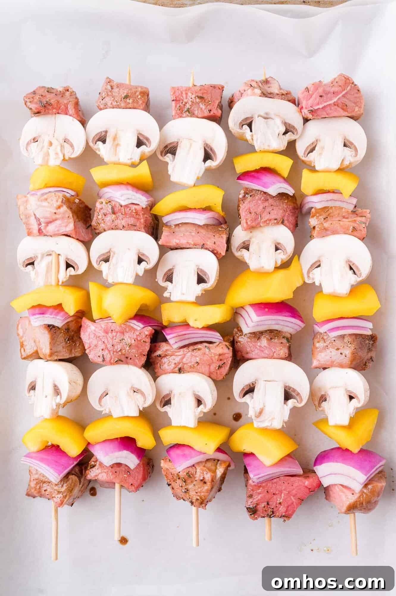 Five skewers with alternating pieces of raw beef, white mushrooms, yellow bell peppers, and red onions on parchment paper.