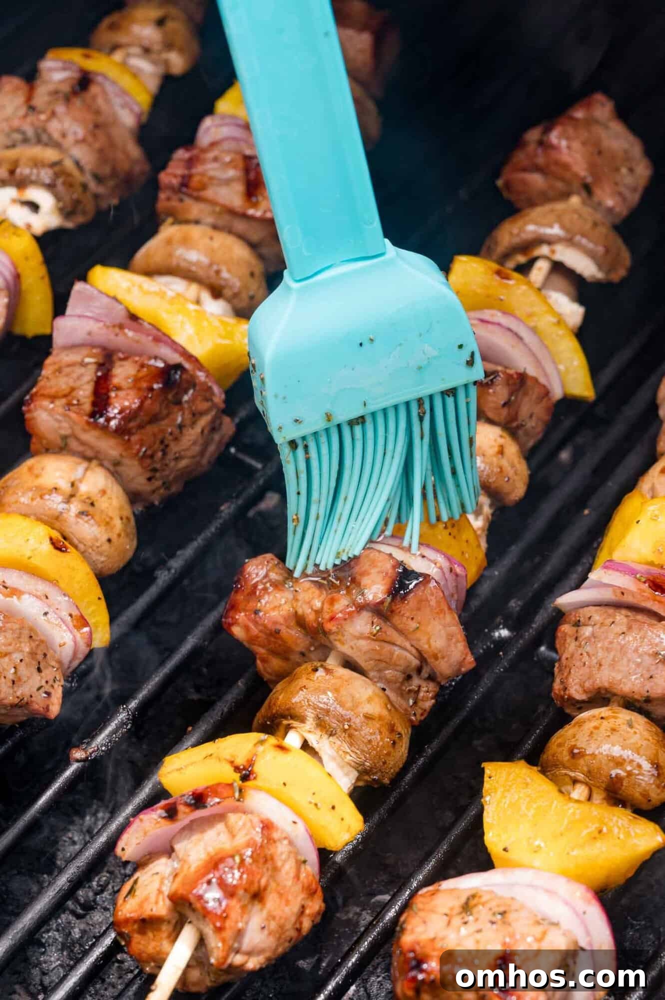 A silicone brush applies marinade to meat and vegetable skewers grilling on a barbecue.