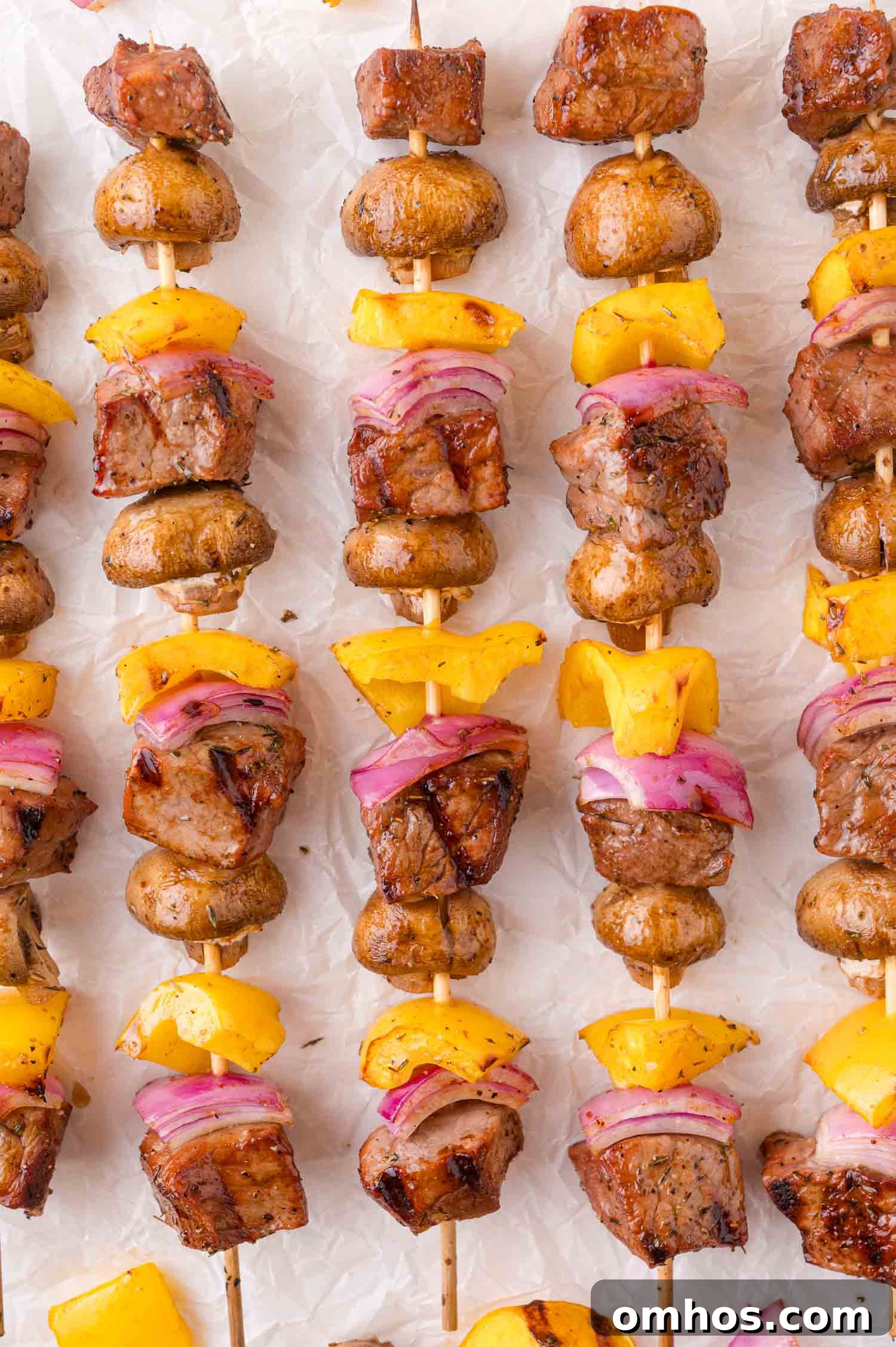 Skewers with grilled beef, mushrooms, yellow bell peppers, and red onions arranged in rows on a white surface.