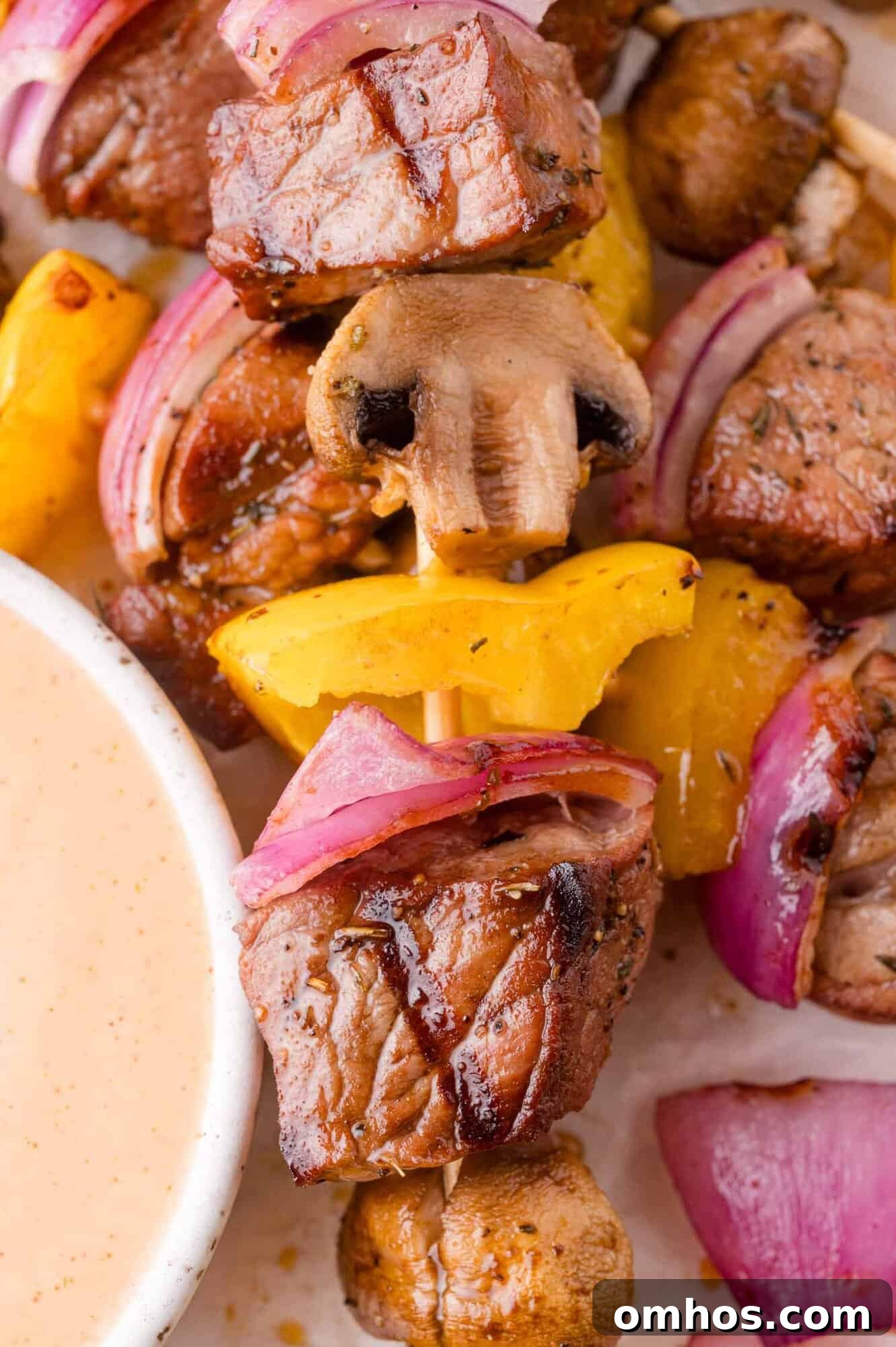 Close-up of a skewer with grilled beef, yellow bell pepper, red onion, and mushroom next to a bowl of dipping sauce.