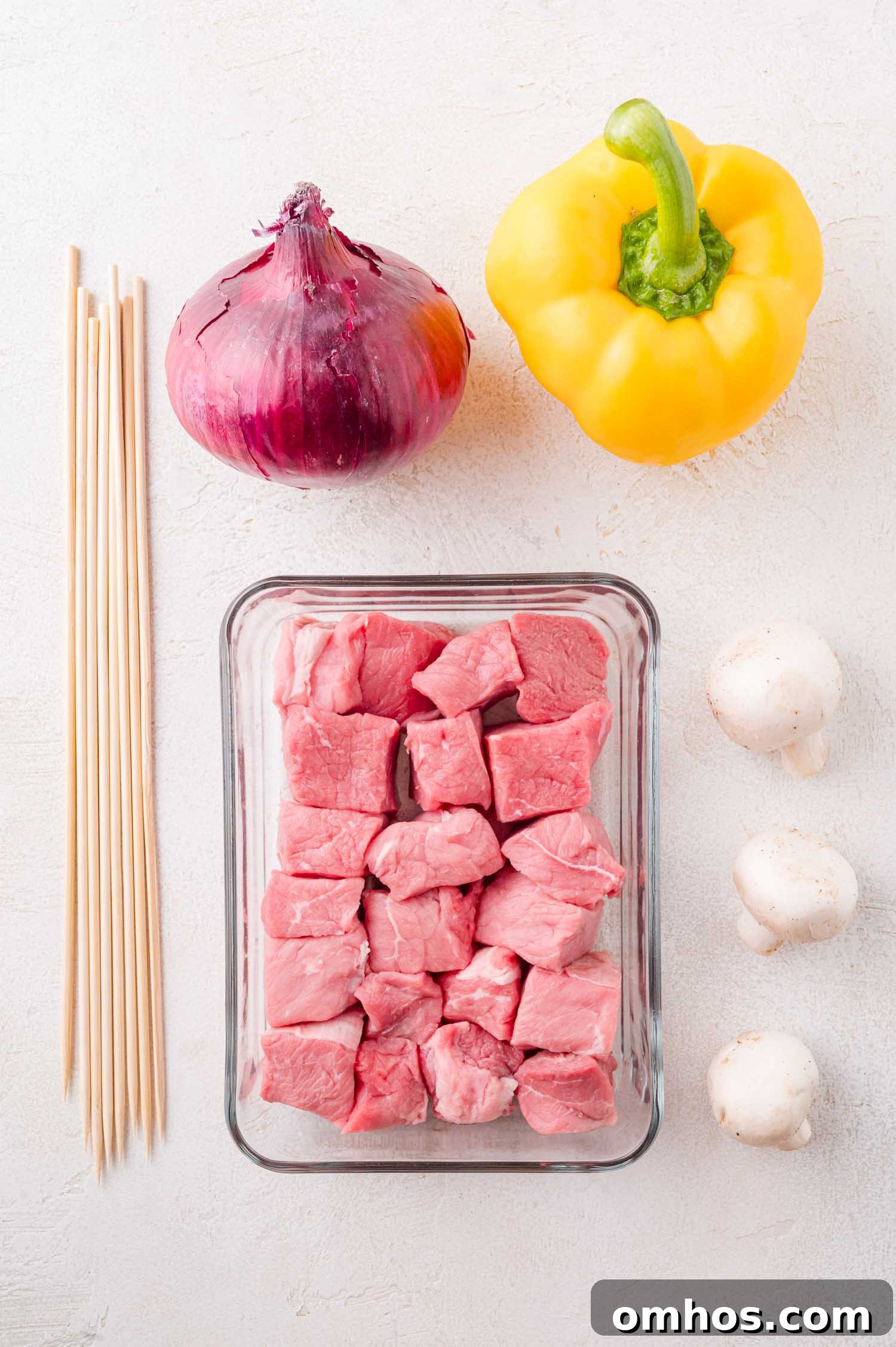 Glass container with raw steak cubes, a red onion, yellow bell pepper, two white mushrooms, and wooden skewers arranged on a white surface.