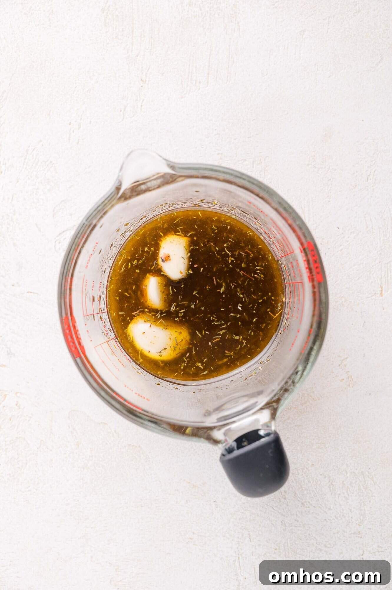 A glass measuring cup containing brown liquid with herbs and two cloves of garlic on a light surface.