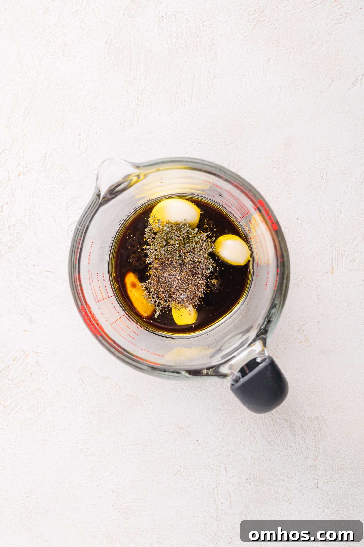 A glass measuring cup filled with dark liquid, three garlic cloves, and herbs, viewed from above on a light textured surface.