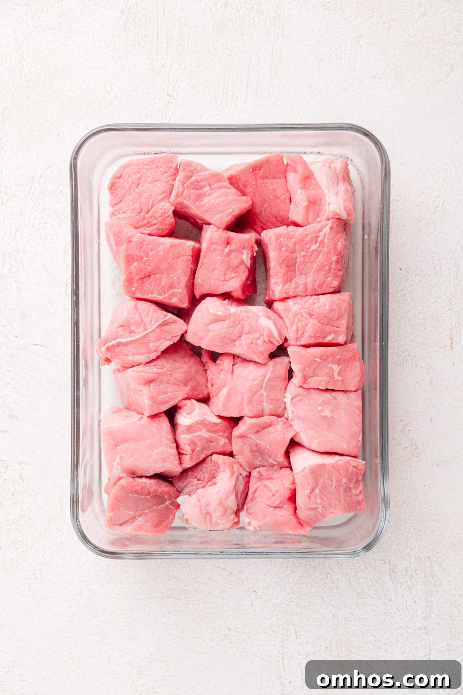 Glass container filled with raw, cubed pieces of beef on a white surface.