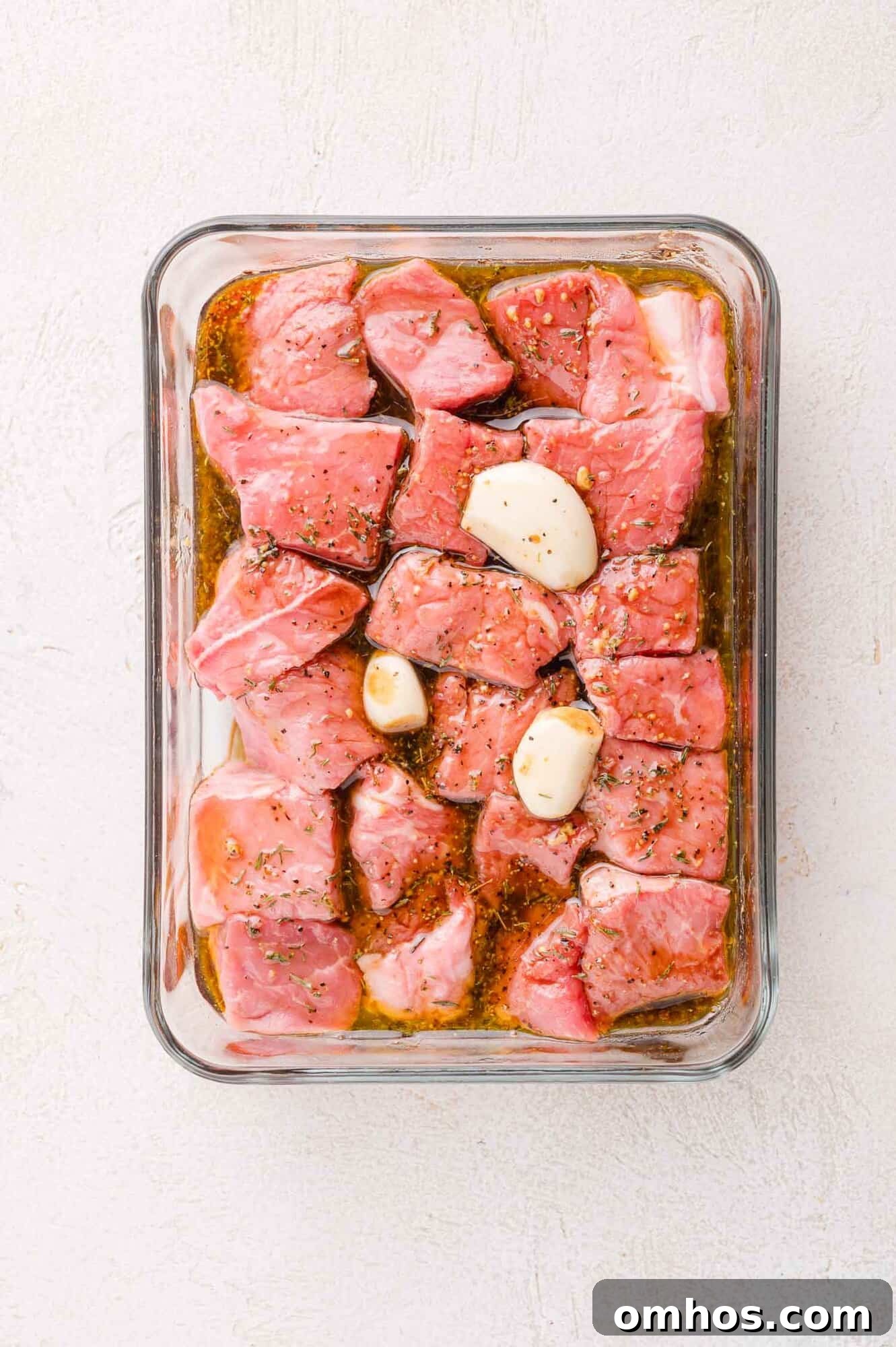 Raw beef cubes and whole garlic cloves marinating in a rectangular glass dish with herbs and seasoning on a light background.