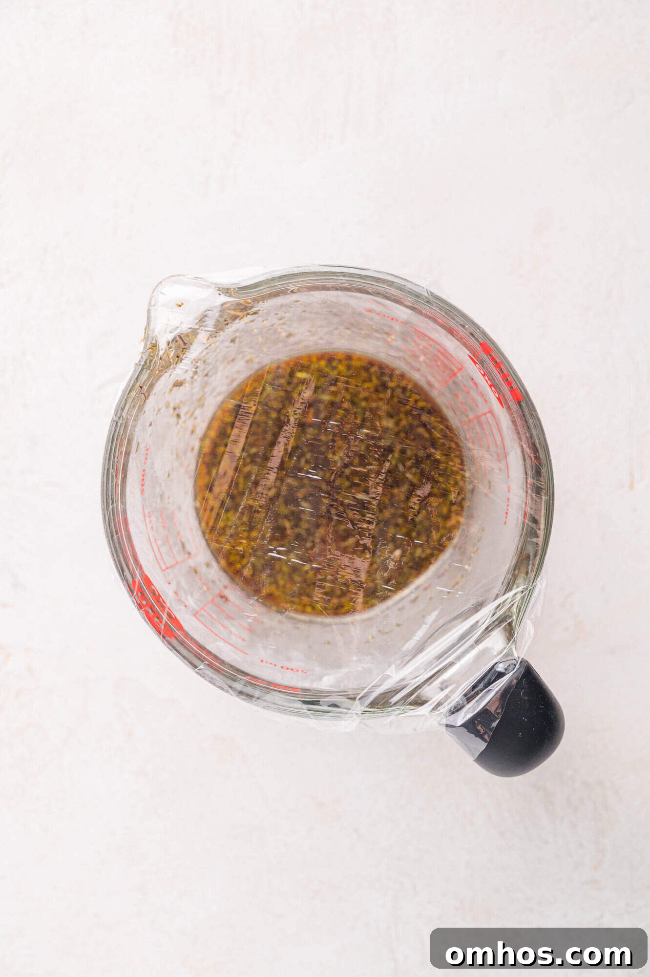 A glass measuring cup filled with brown liquid, covered with plastic wrap, placed on a white surface.