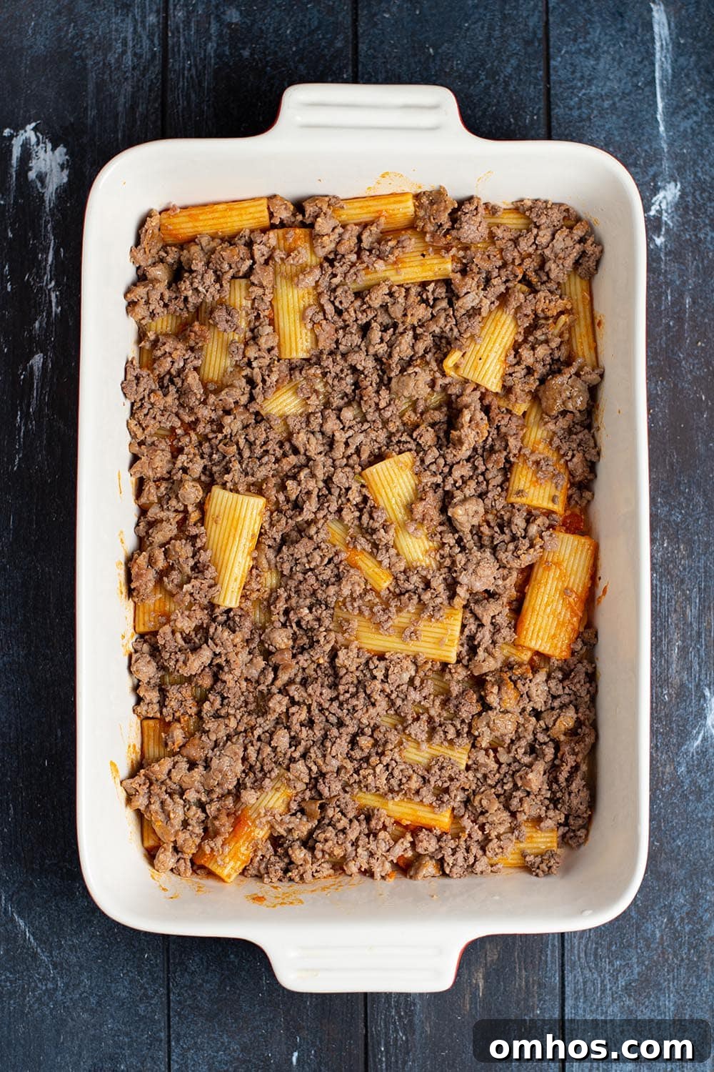 A layer of cooked ground beef and sausage on top of rigatoni noodles in a casserole dish.