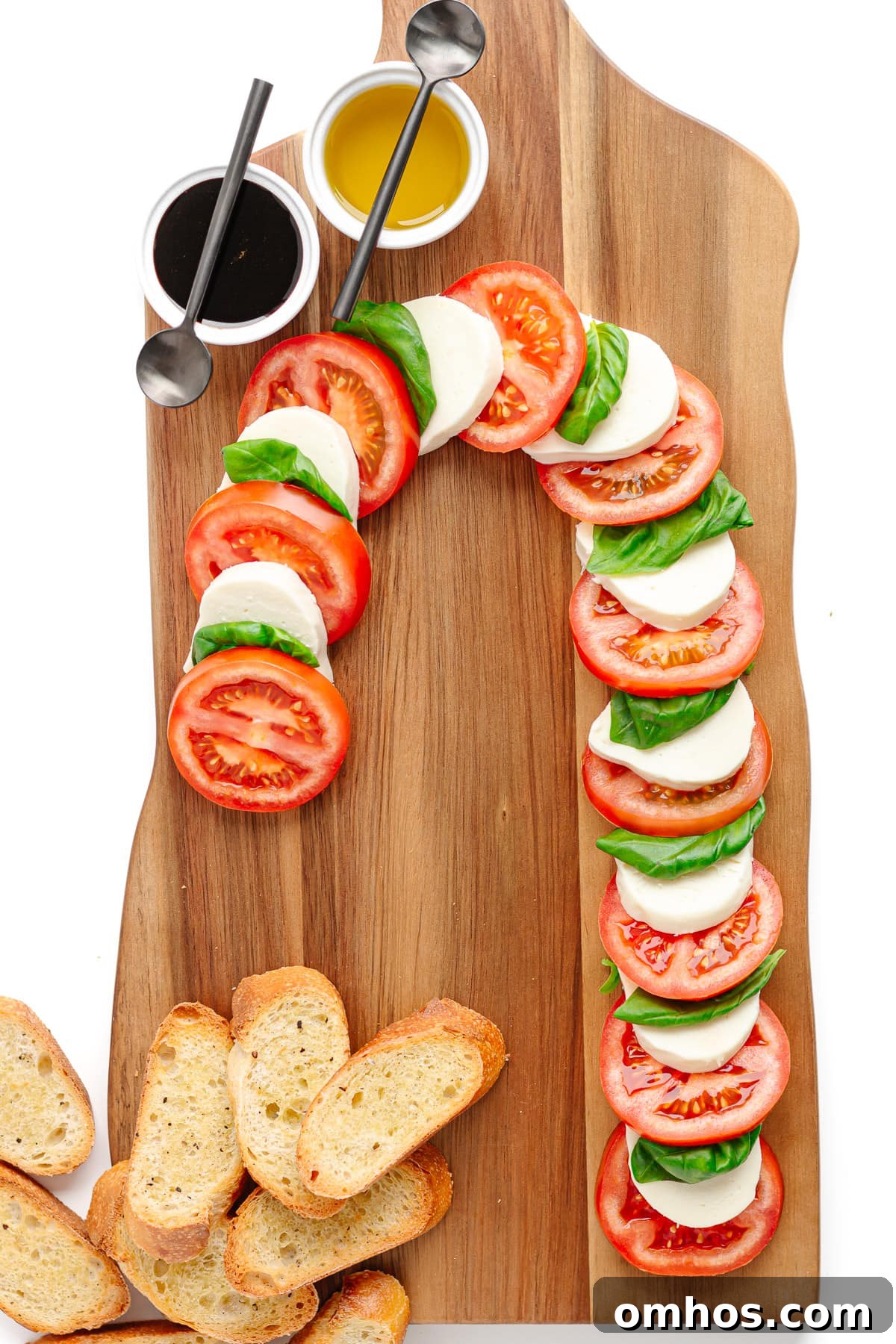 Tomato slices, fresh mozzarella slices and basil leaves arranged into a candy cane shape on a wooden board along with crostini and two small bowls containing balsamic glaze and olive oil.