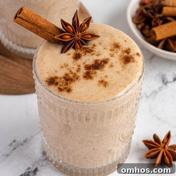Close up of a light brown banana chai spice smoothie in a glass garnished with a cinnamon stick, star anise, and a dusting of ground spices.