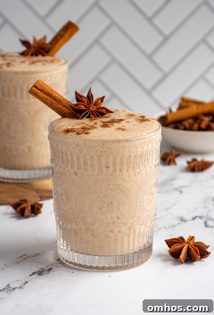 A glass filled with light brown banana chai smoothie garnished with a cinnamon stick, star anise, and a dusting of ground spices.