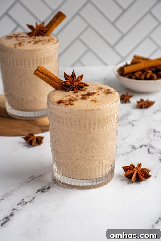 Glass cup filled with light brown banana chai smoothie garnished with cinnamon stick, star anise, and a dusting of spices.