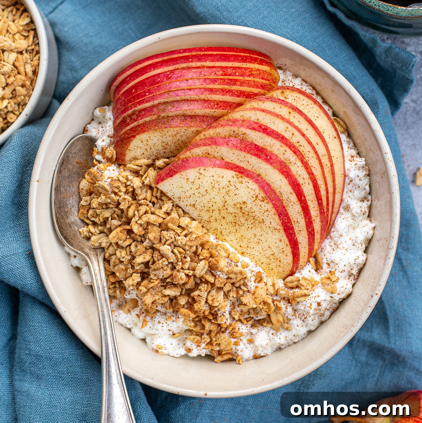 A white bowl on a blue towel filled with cottage cheese and apples with granola and ground cinnamon.