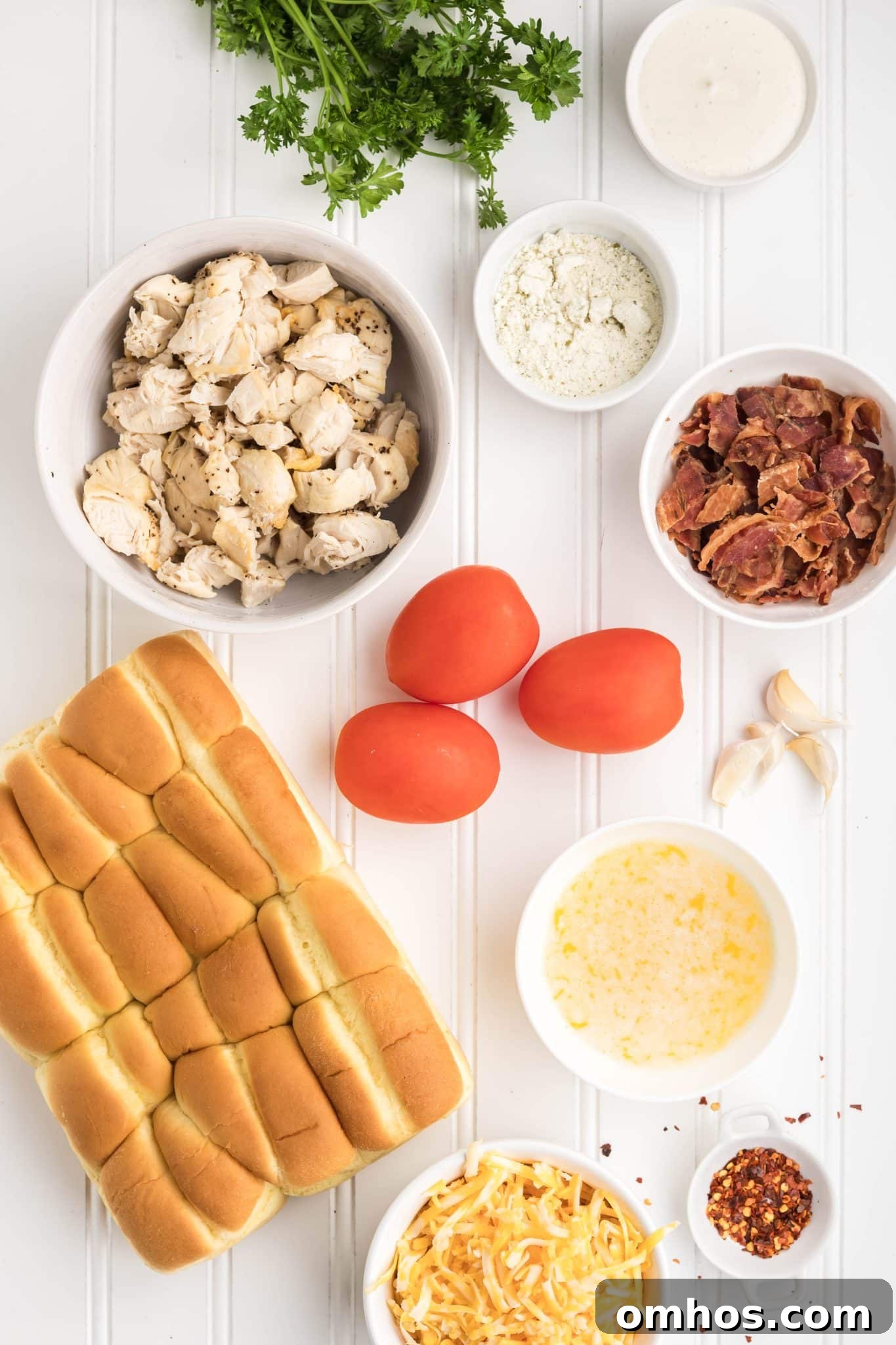 Ingredients for chicken bacon ranch sliders arranged on counter including Hawaiian rolls, chopped chicken, crispy bacon, ranch dressing, shredded cheese, red chili flakes, and fresh tomatoes.