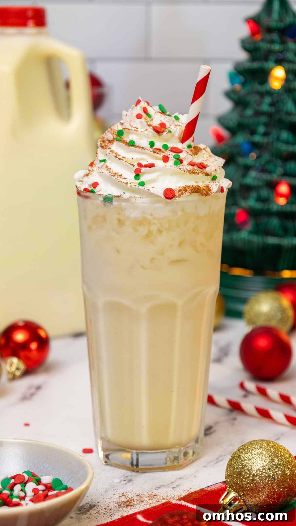 Eggnog Dirty Soda: Holiday Cocktail Recipe 2 Tall glass of eggnog dirty soda topped with whipped cream, red and green holiday sprinkles, and a dusting of cinnamon, served with a red-striped straw. The festive background includes Christmas ornaments, a ceramic Christmas tree, and a carton of eggnog.