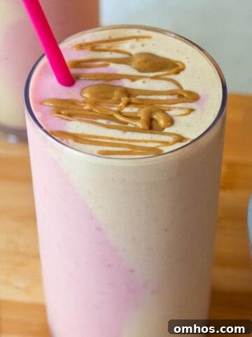 A tall glass of peanut butter and jelly smoothie with pink and beige side-by-side in the glass, topped with peanut butter drizzle.