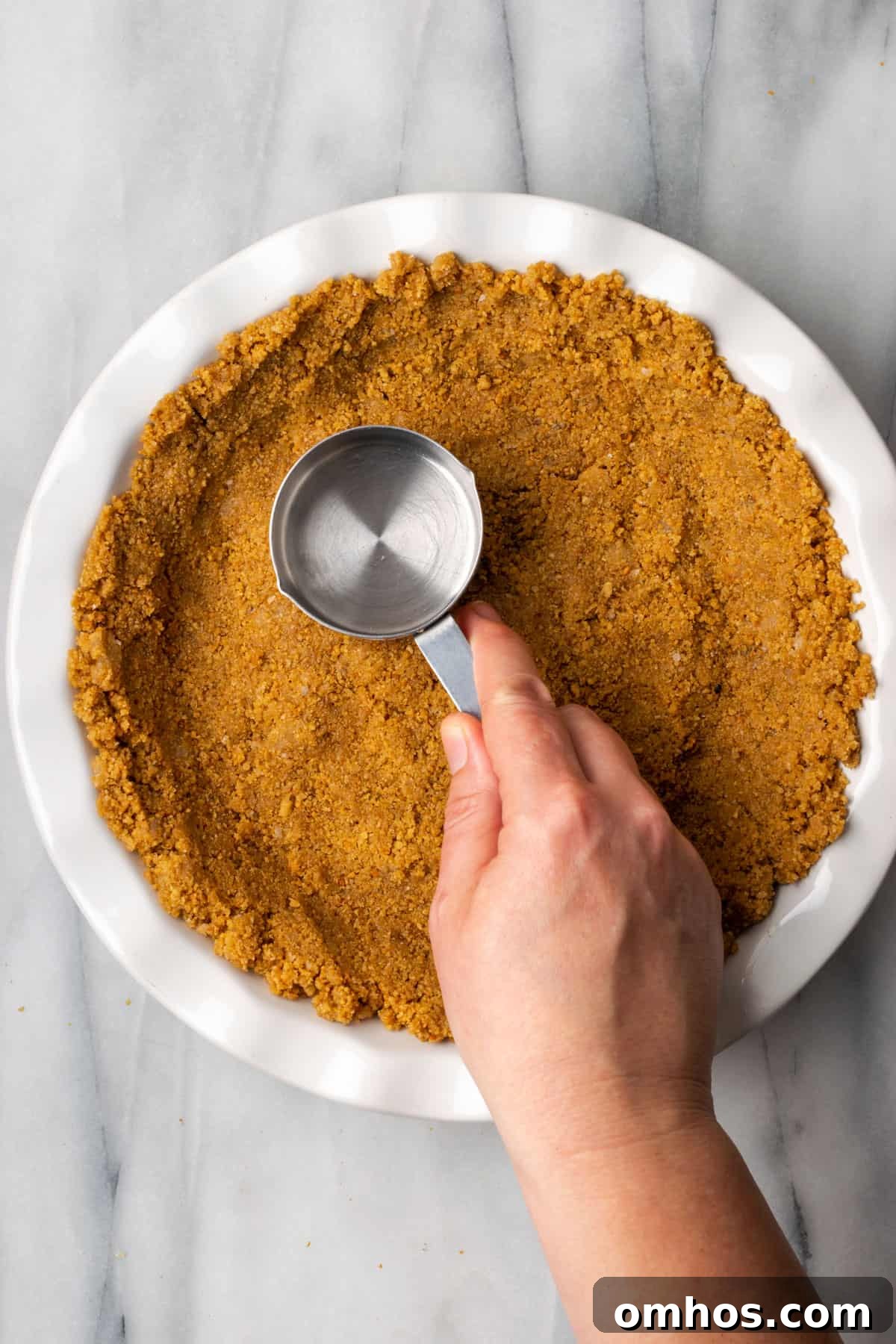 A hand pressing graham cracker crust mixture into a pie plate with a measuring cup. 