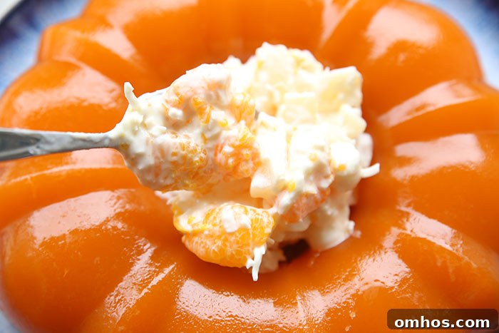A scoop of ambrosia topping filling the center hole of a bundt-shaped orange Jello salad on a round serving plate.
