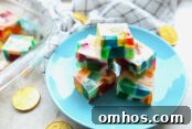 Broken Glass Jello Squares stacked on a small blue plate. Surround the plate are Euro coins. In the upper background is the corner of a glass dish with jello squares and a kitchen towel.