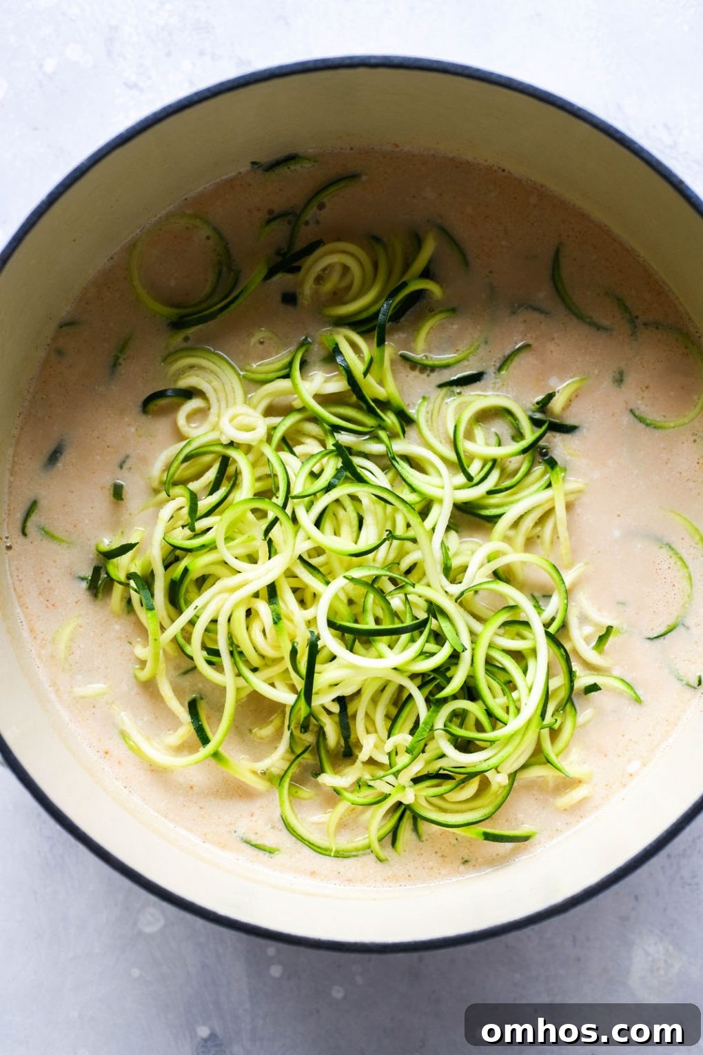 Overhead shot of a large white ceramic soup pot filled with coconut broth, and bright green raw zucchini noodles.