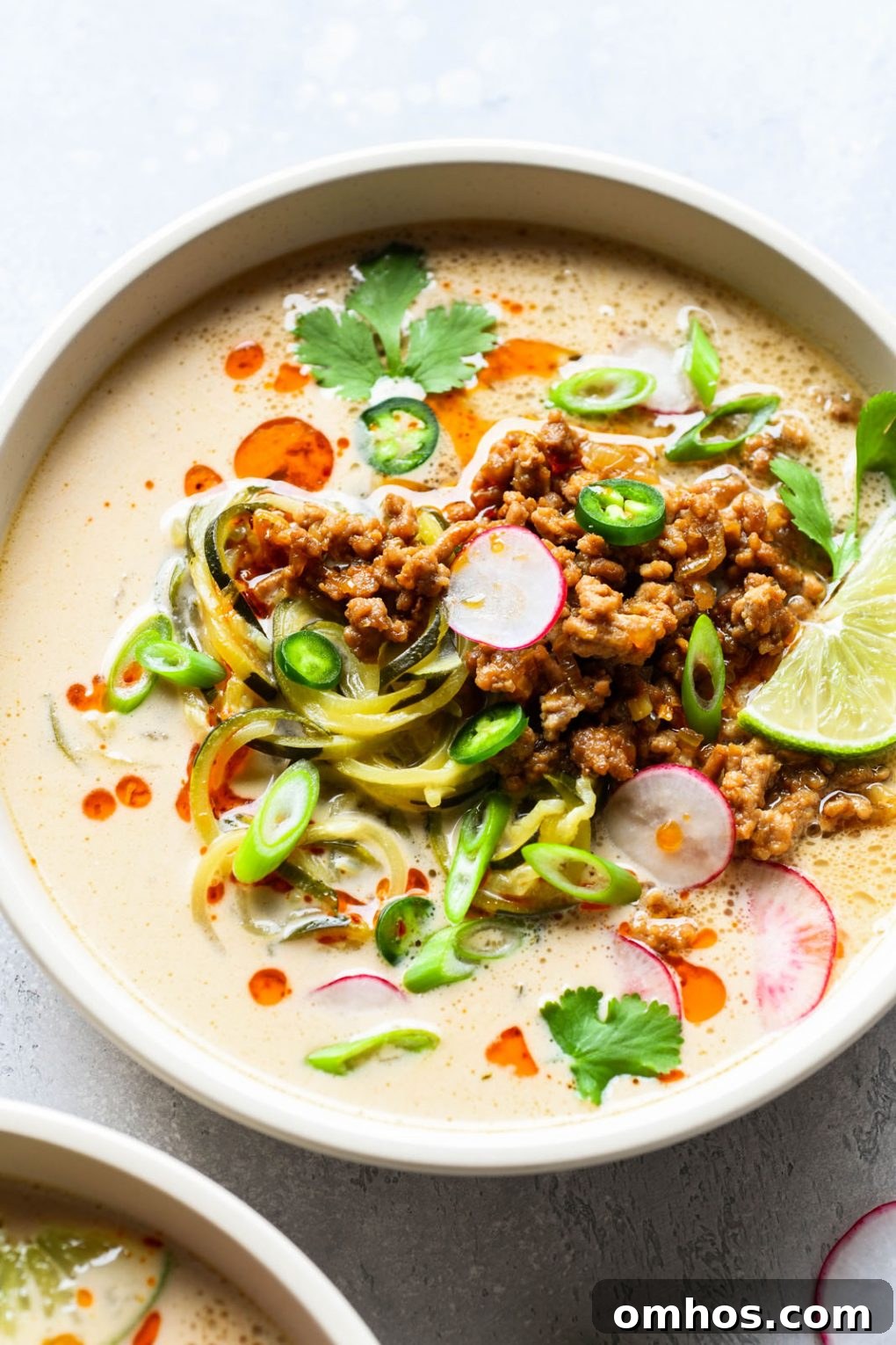 Overhead shot of a bowl of zucchini noodle soup with crispy pork and a coconut broth. Topped with thinly sliced radishes, green onions, jalapeno, cilantro, chili oil, and lime wedges. In a light colored bowl on a white background.