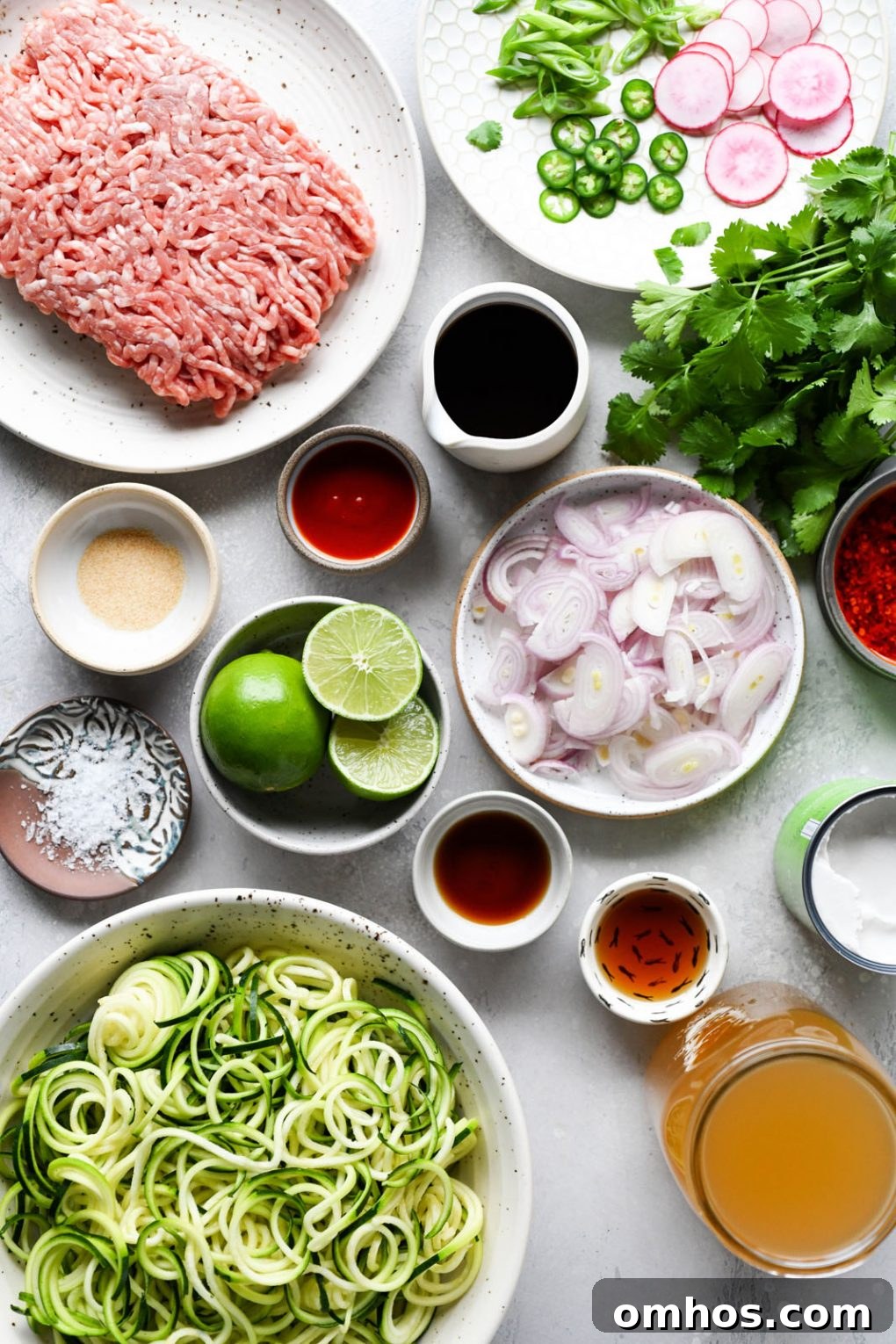 Overhead shot of the different ingredients needed to make this crispy pork and zucchini noodle coconut soup. Ground pork, thinly sliced shallots, zoodles, chicken broth, spices, limes, cilantro, and various sauces in small bowls. On a light colored background.