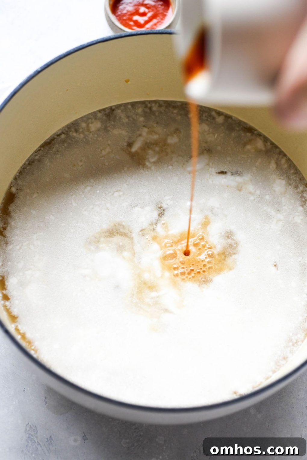 Large white ceramic pot with coconut broth, with a hand pouring coconut aminos into the pot.