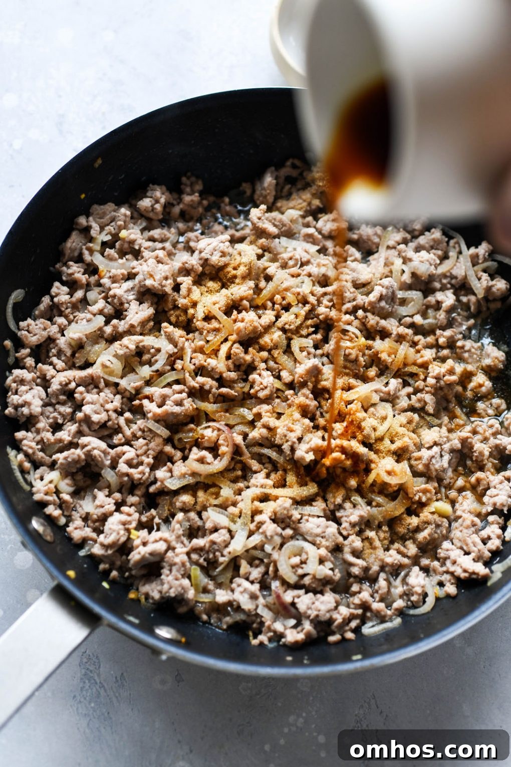 Close up shot of cooked ground pork, with a hand pouring coconut aminos into the pan.