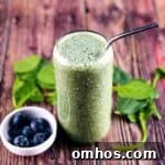 A delicious spinach smoothie poured into a tall glass, with a small bowl of blueberries beside it, and spinach leaves behind it.