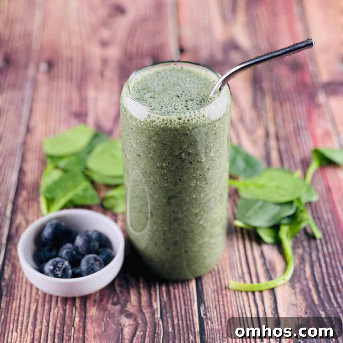 Spinach Banana Smoothie Recipe 6 A creamy spinach smoothie in a tall glass with a straw, with a bowl of blueberries and spinach leaves.
