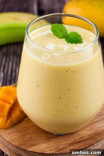 A small glass of healthy mango banana smoothie garnished with a mint leaf and a fresh mango and banana beside it.