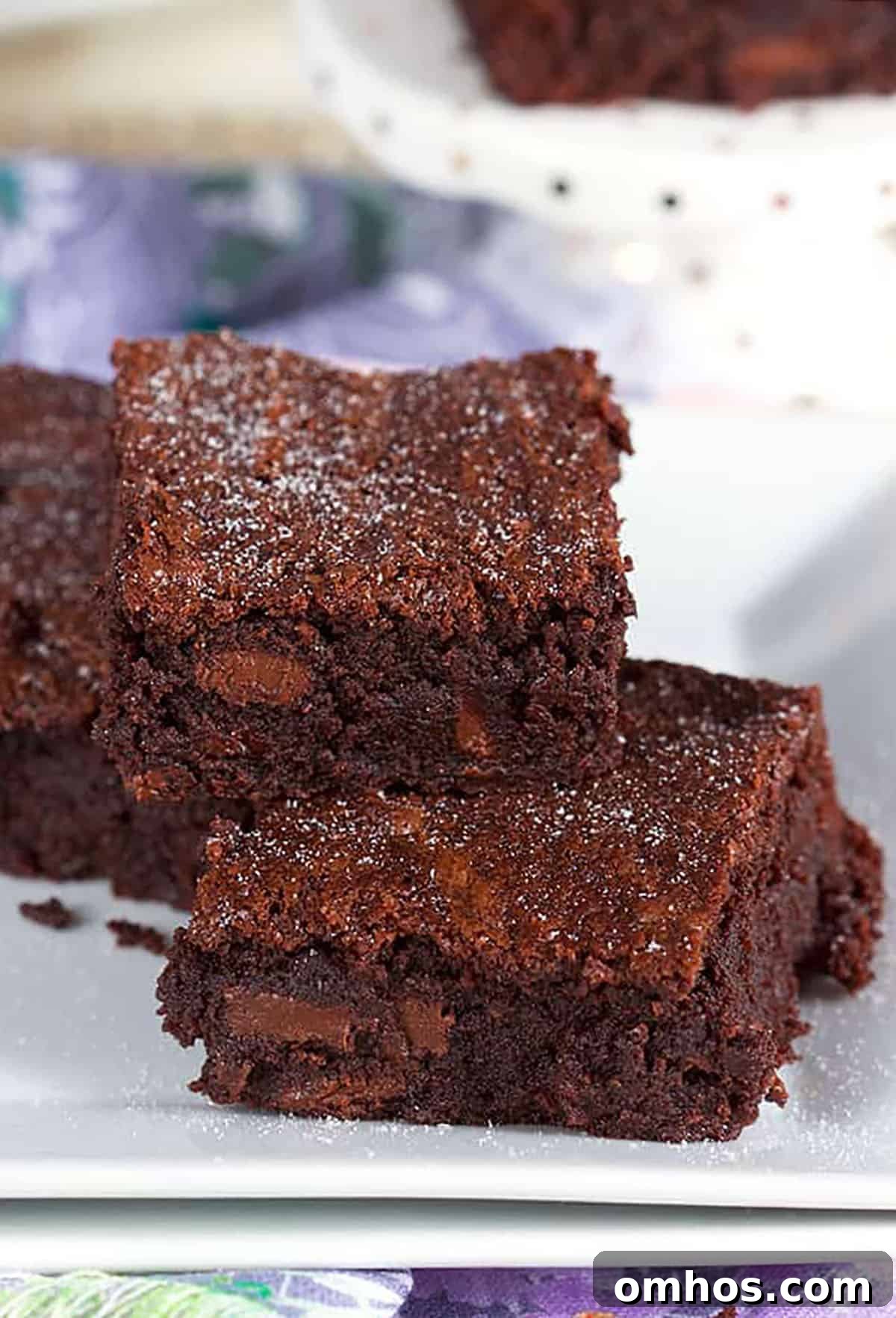 Three brownies piled on a white plate with confectioners sugar sprinkles on top.