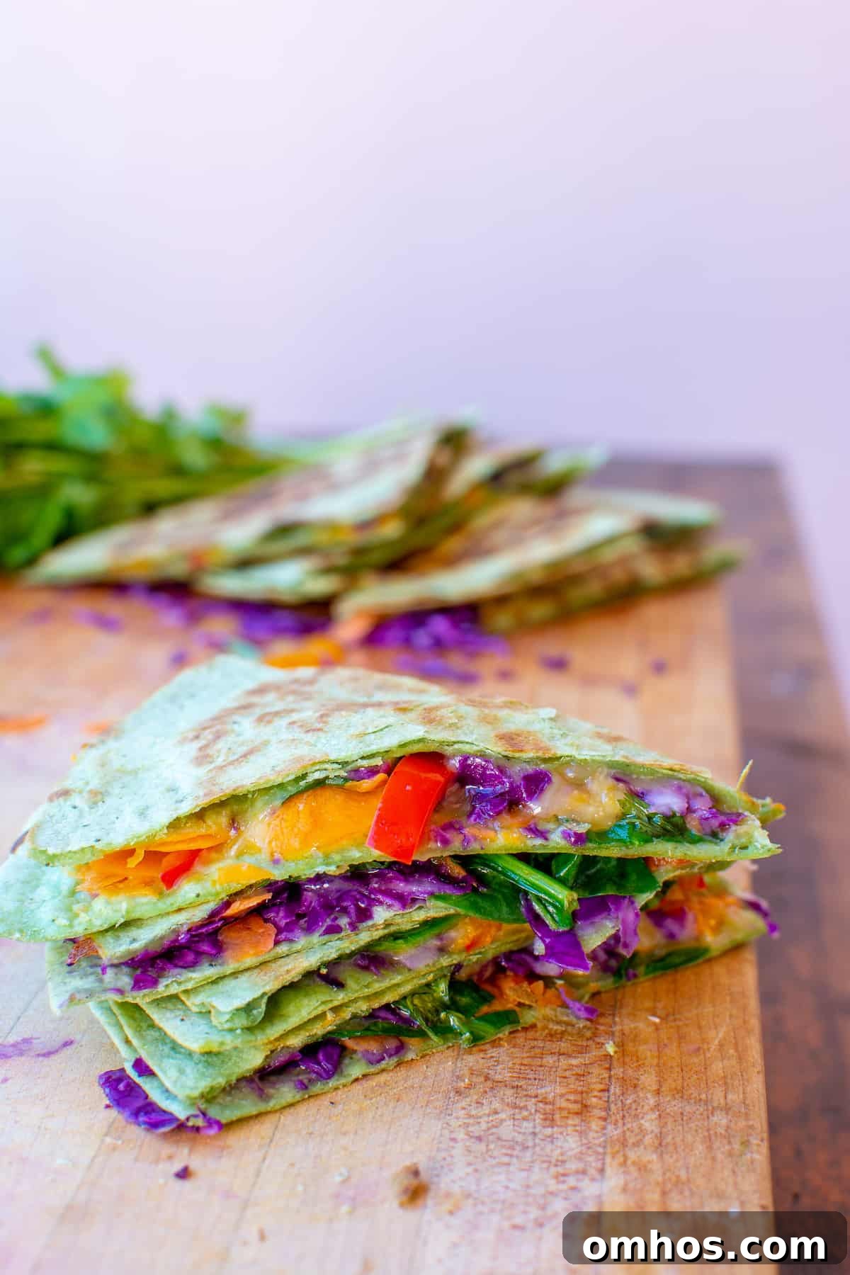 How to Make a Rainbow Vegetable Quesadilla 6 A stack of vegetable quesadilla slices sitting on a cutting board on top of a wood table with bell peppers, red cabbage, and cheese.