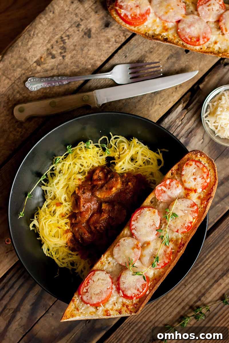 Spaghetti Squash Marinara with Chili Oil Tomato Garlic Bread 7 Vertical top-down image of a bowl of squash with marinara, with bread topped with tomatoes and melted cheese on the side, with a metal fork and knife on a wooden surface.