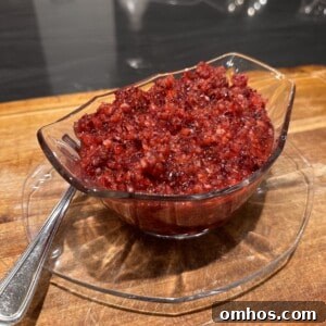 A glass bowl filled with a chunky, red cranberry relish sits on a matching glass plate atop a wooden surface. A metal spoon is placed beside the bowl.