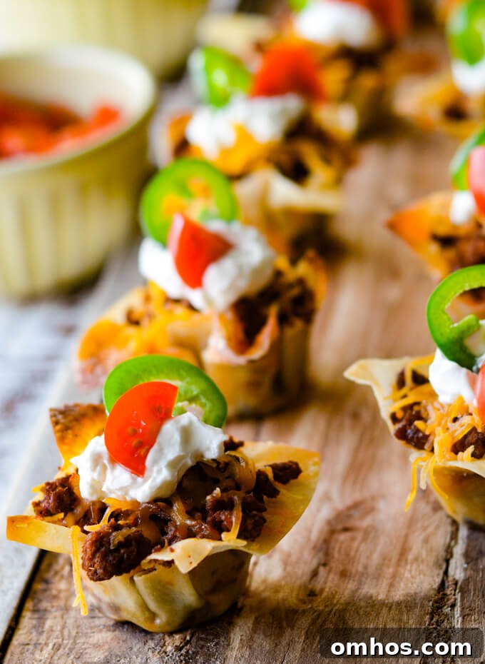 Mini Taco Wonton Cups with sour cream, cherry tomato and jalapeno slices. All of these appetizers are sitting on a wooden board with a cup of tomatoes sitting next to it.