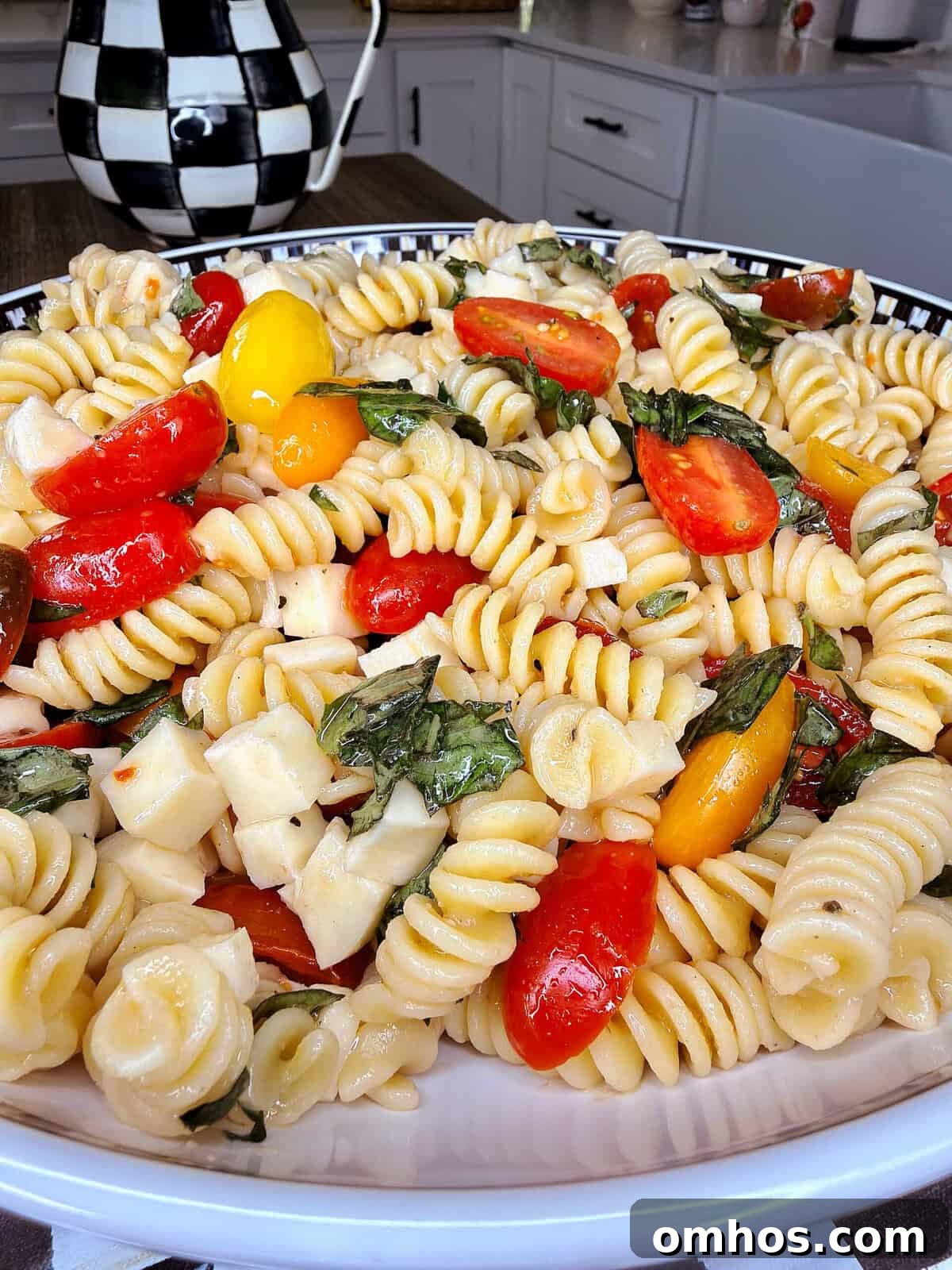 A close-up of an Easy Italian Pasta Salad with rotini, cherry tomatoes, basil leaves, and mozzarella chunks served in a decorative bowl in a kitchen setting.
