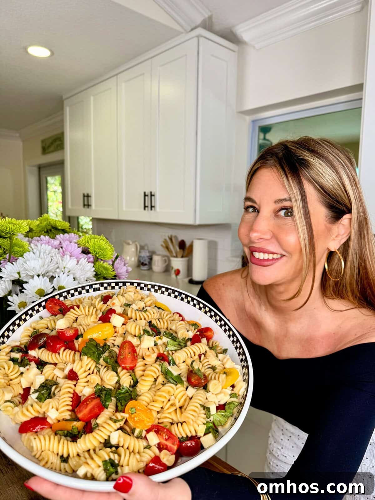 A woman smiles while holding a large bowl of Easy Italian Pasta Salad with tomatoes, basil, and cheese in a bright kitchen.