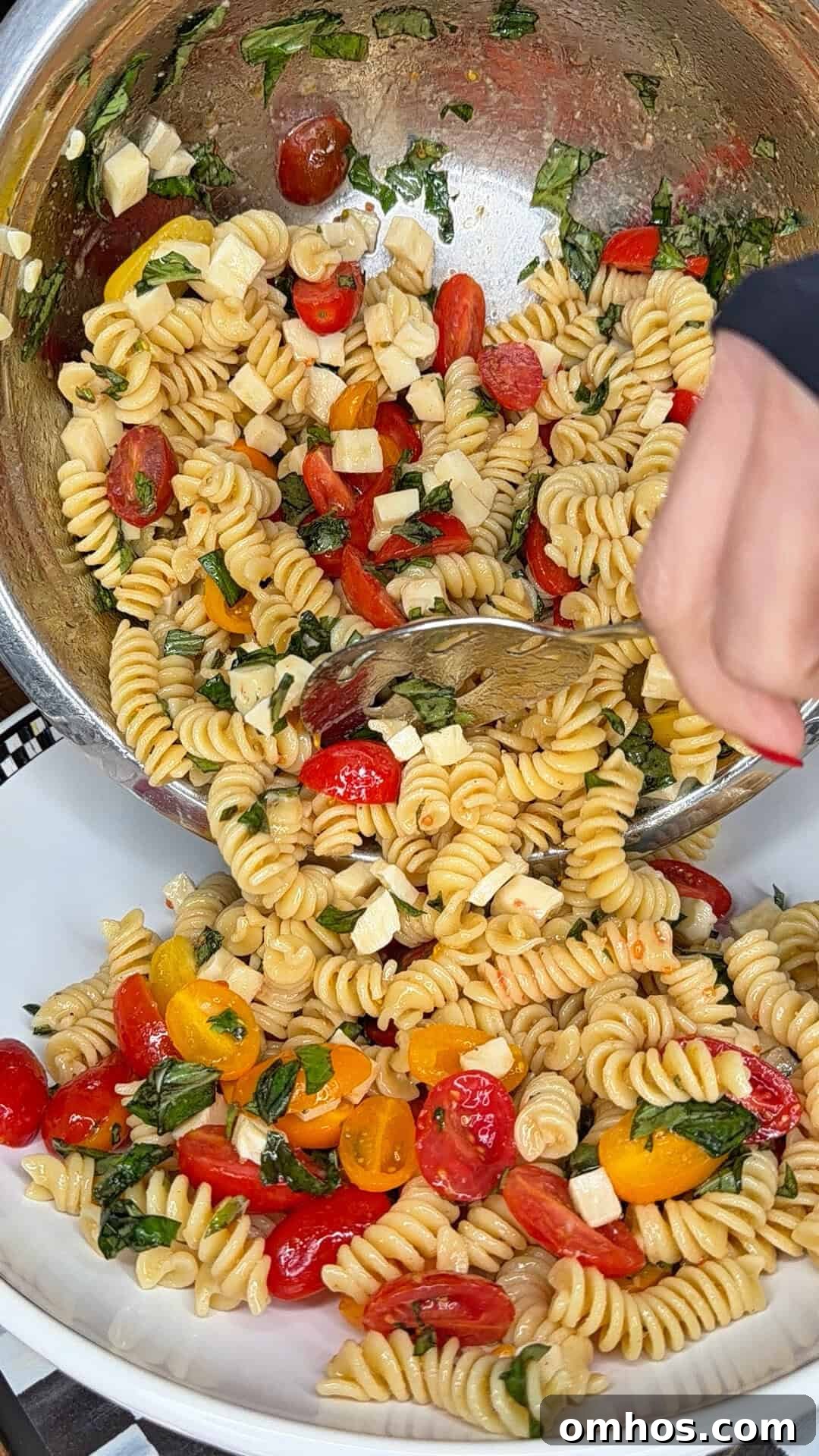 A hand serves Easy Italian Pasta Salad—rotini with cherry tomatoes, mozzarella cubes, and fresh basil—from a large metal mixing bowl into a white dish.