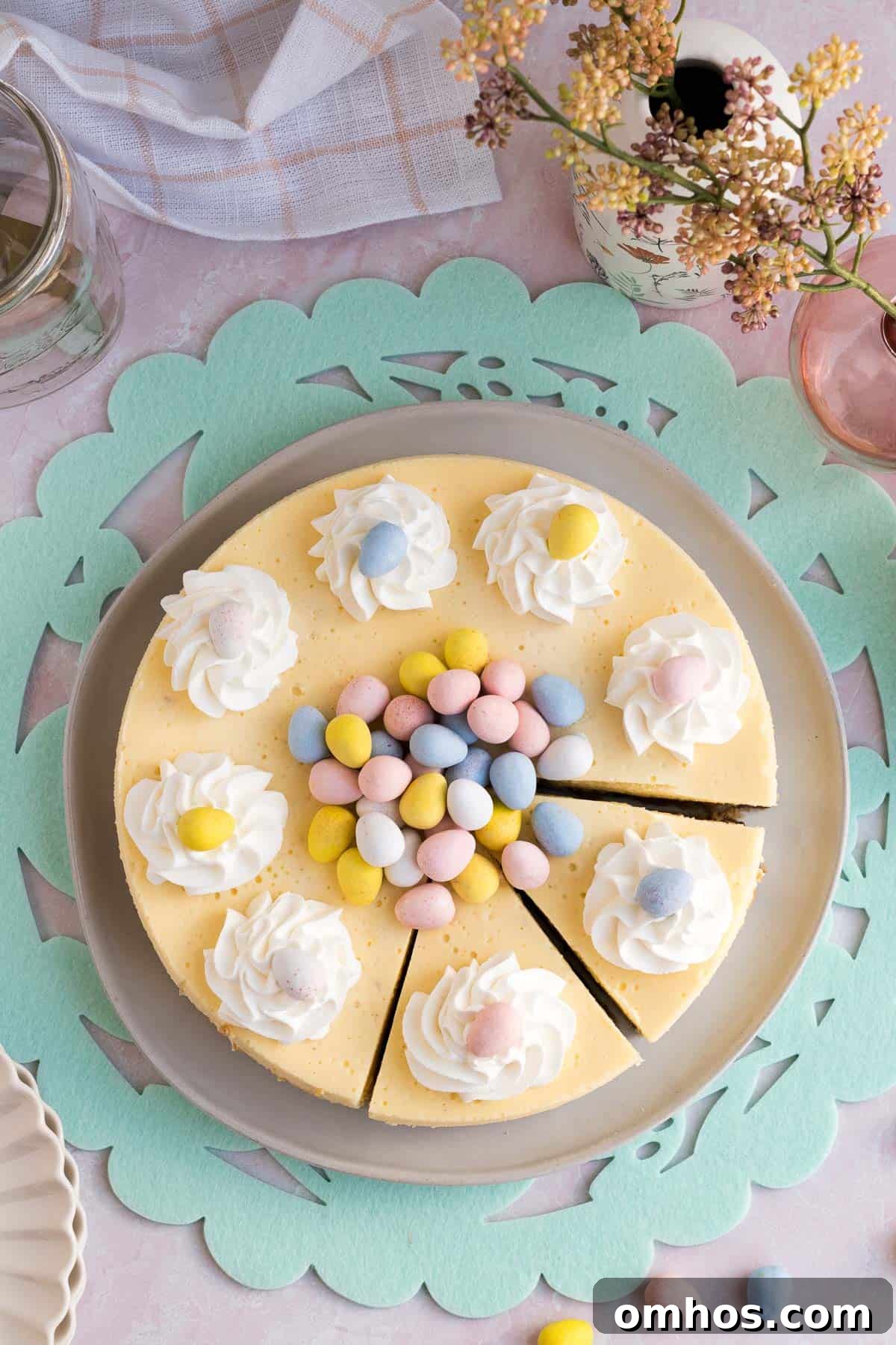 No-Bake Easter Egg Cheesecake Recipe 2 Top view of an Easter egg cheesecake with whipped cream and mini eggs piled on top and 2 slices cut sitting on a green Easter placemat