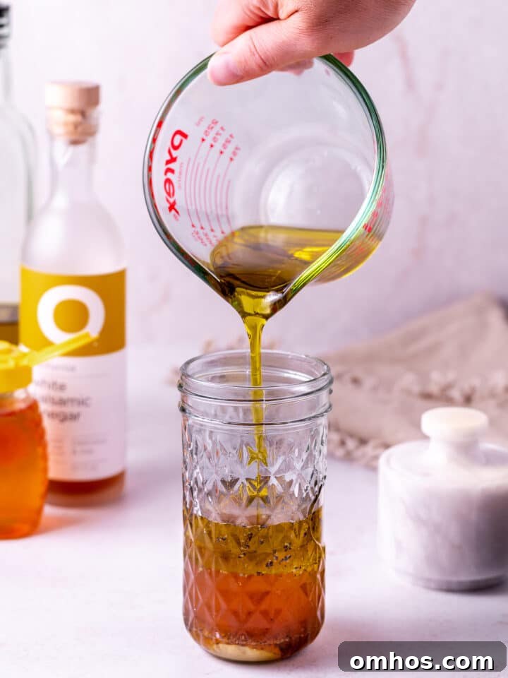 hand pouring olive oil from a liquid measuring cup into a mason jar with other salad dressing ingredients