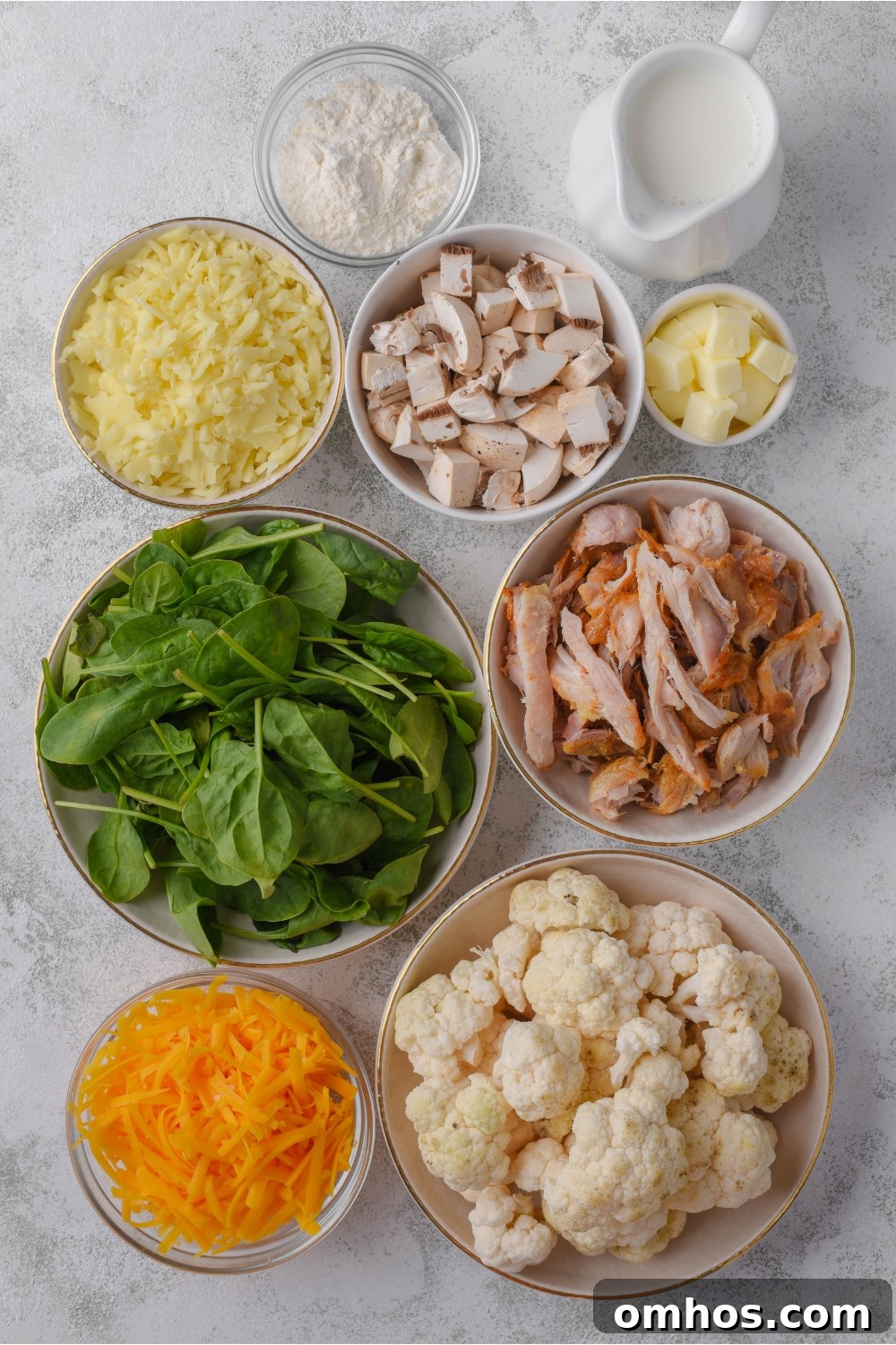 An assortment of ingredients including bowls of fresh spinach, shredded chicken, diced mushrooms, cauliflower florets, shredded cheese, flour, and butter cubes.