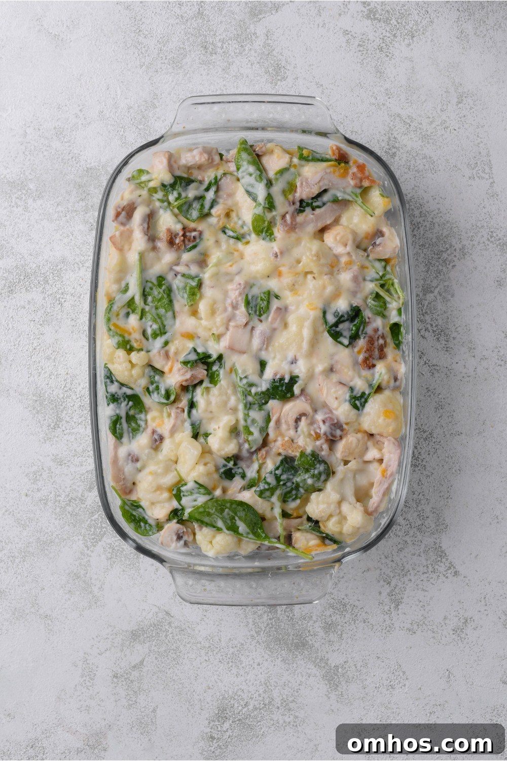 A baking dish filled with unbaked chicken, cauliflower and spinach casserole.