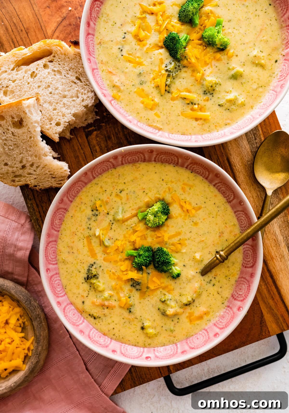 Broccoli cheddar soup recipe in a pink bowl with a spoon in it ready to serve. crusty bread on the side. Two bowls ready to eat.